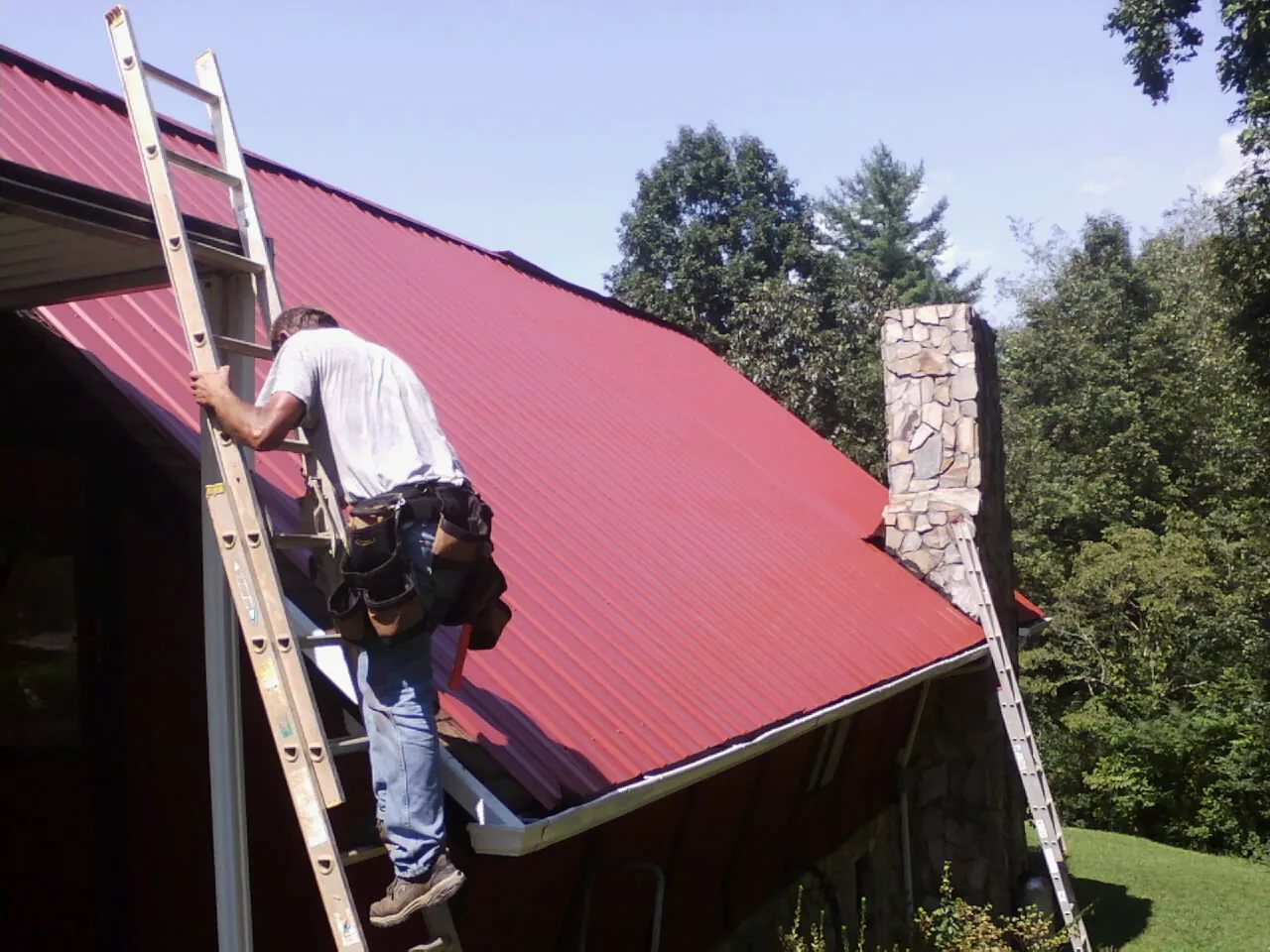 Roofer on a ladder beside a red metal roof and stone chimney — Soffit & Fascia Repair by Roofing Brothers in Papillion